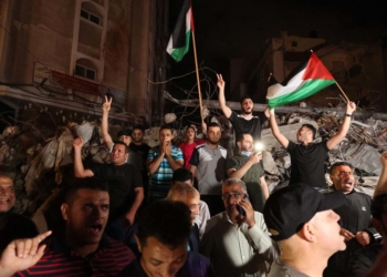 A man waves the Palestinian flag as others flash the V-sign for victory as they celebrates in front of a destroyed building the ceasefire brokered by Egypt between Israel and the two main Palestinian armed groups in Gaza on May 21, 2021. - Israel and the two main Palestinian armed groups in Gaza, Hamas and Islamic Jihad, announced a ceasefire on May 20, 2021, aimed to end the most devastating conflict between them for seven years. The truce brokered by Egypt was announced following mounting international pressure to end 11 days of conflict that has claimed lives on both sides, with Israeli jets pounding Gaza with air strikes as militants fired thousands of rockets towards Israel. (Photo by MOHAMMED ABED / AFP)